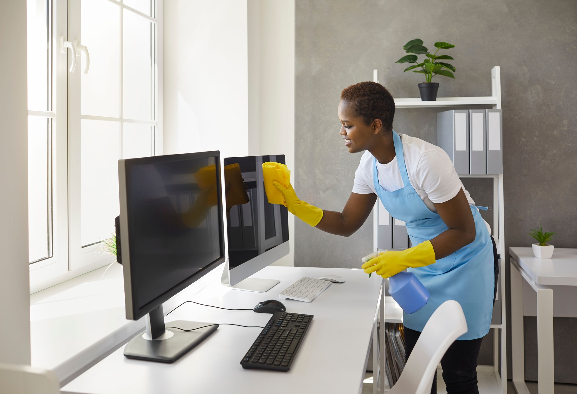 Happy woman from janitorial cleaning service wiping computer screens in modern office